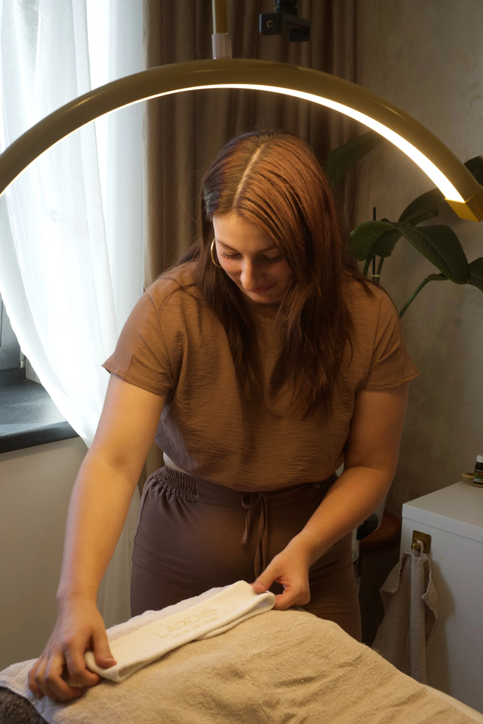 Aneta ve studiu Valany rozkládá ručník LARENS na kosmetickém lehátku, nad ní svítí mosazná kruhová lampa, za ní záclona a monstera.